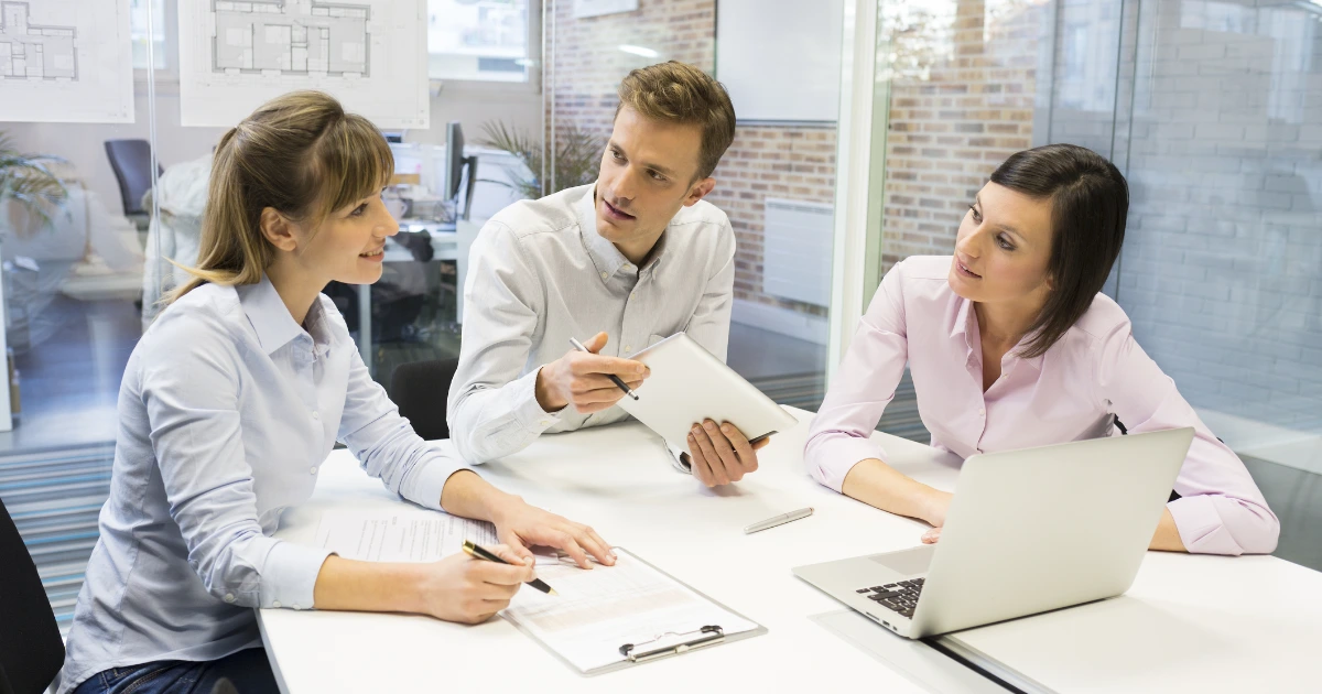 Three colleagues in business-casual attire discuss notes and a laptop at a white meeting table in a bright office.