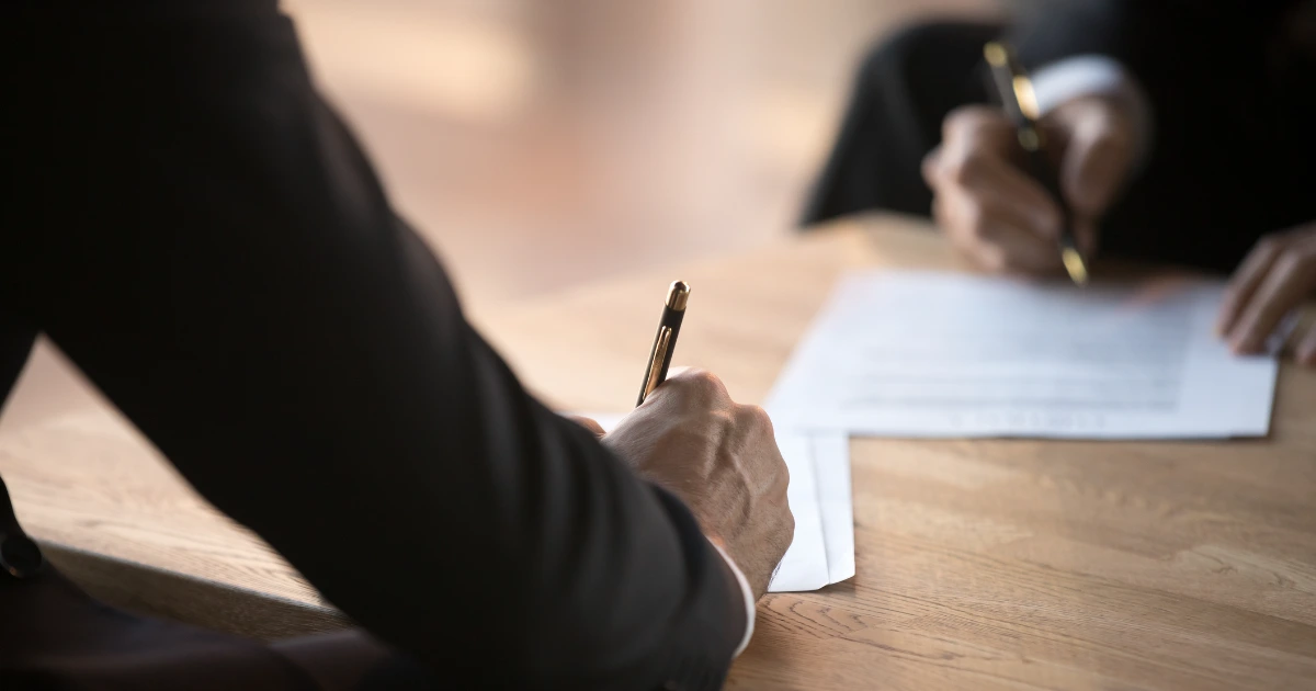 Two suited individuals signing printed documents with gold pens on a wooden table, close-up shot.