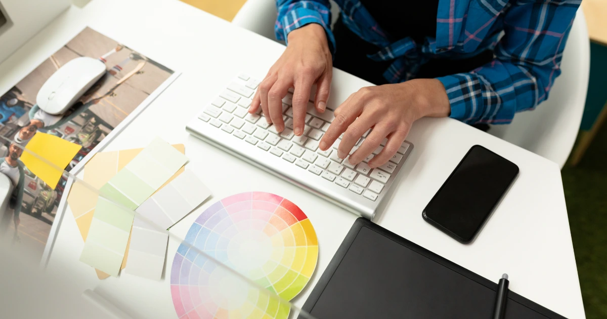 Person in plaid shirt typing at keyboard surrounded by colour swatches, graphics tablet, phone and printed photos