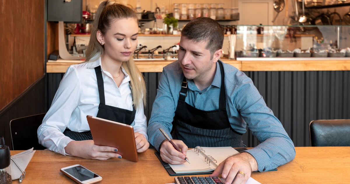 Two café owners in black aprons review a tablet and notebook with calculator at a wooden counter