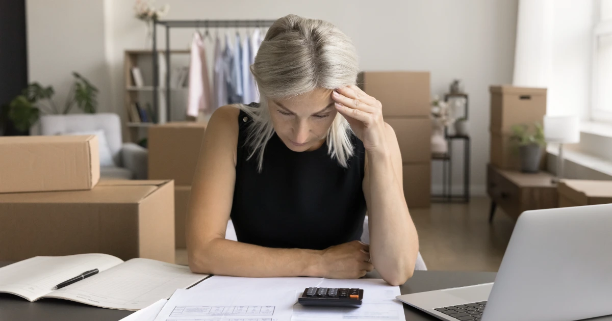 Woman with grey hair studies paperwork and calculator at desk, cardboard boxes and clothing rail behind her