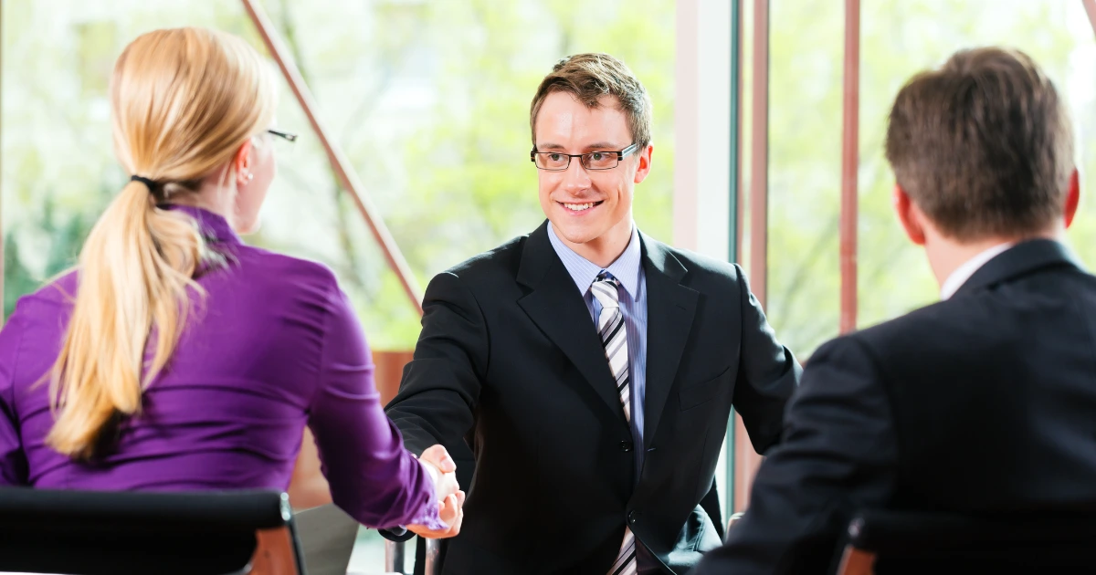 Smiling man in suit and tie shaking hands across a table with two interviewers in a bright office