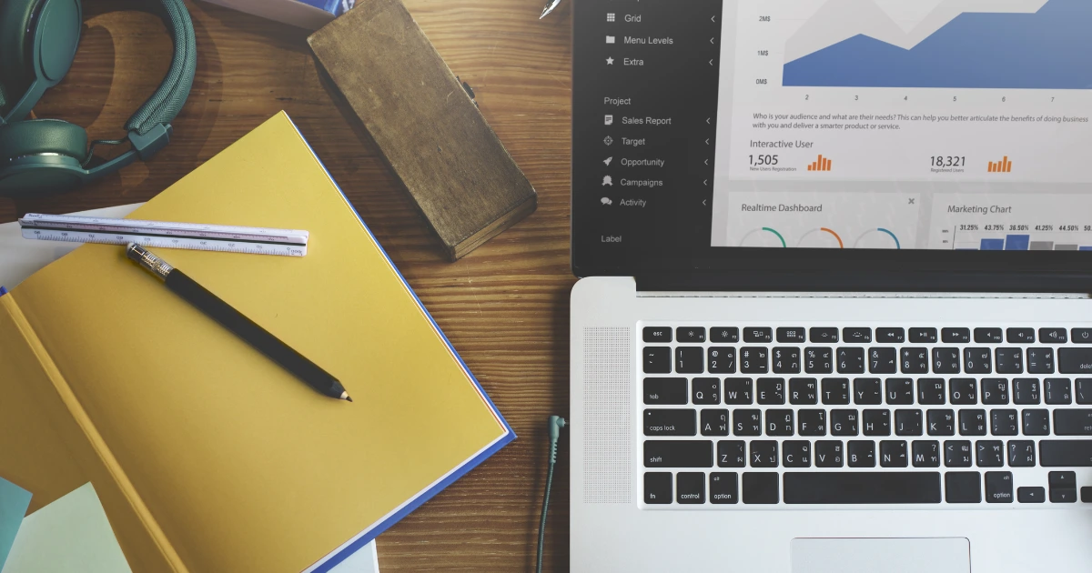 Overhead flatlay of a laptop showing a sales dashboard, yellow notebook, pencil, ruler and headphones on a wooden desk