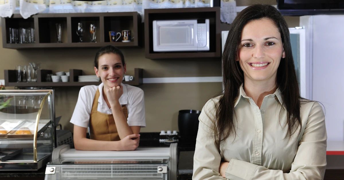 Smiling café owner with arms crossed in foreground, barista in apron leaning on counter behind her