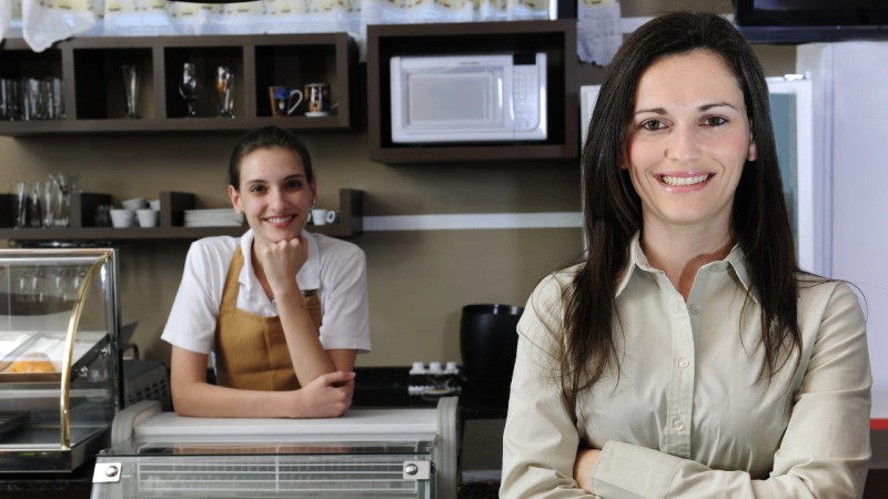 Smiling café owner with arms crossed in foreground, barista in apron leaning on counter behind her