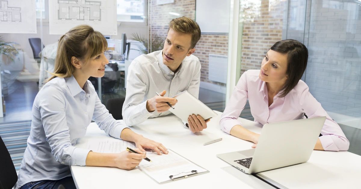 Three colleagues in business-casual attire discuss notes and a laptop at a white meeting table in a bright office.