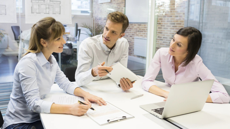 Three colleagues in business-casual attire discuss notes and a laptop at a white meeting table in a bright office.