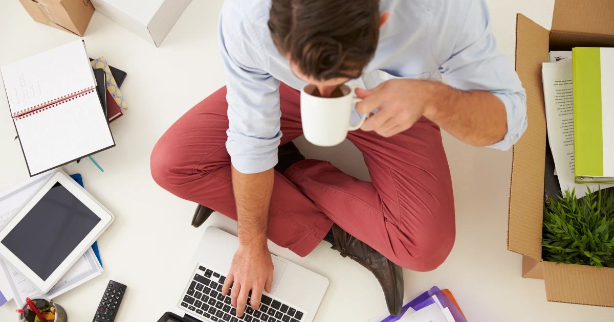 Man sitting cross-legged on floor using laptop and holding a mug, surrounded by notebooks, tablet, and cardboard boxes
