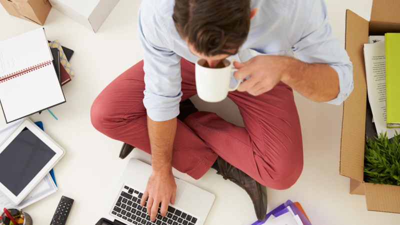 Man sitting cross-legged on floor using laptop and holding a mug, surrounded by notebooks, tablet, and cardboard boxes