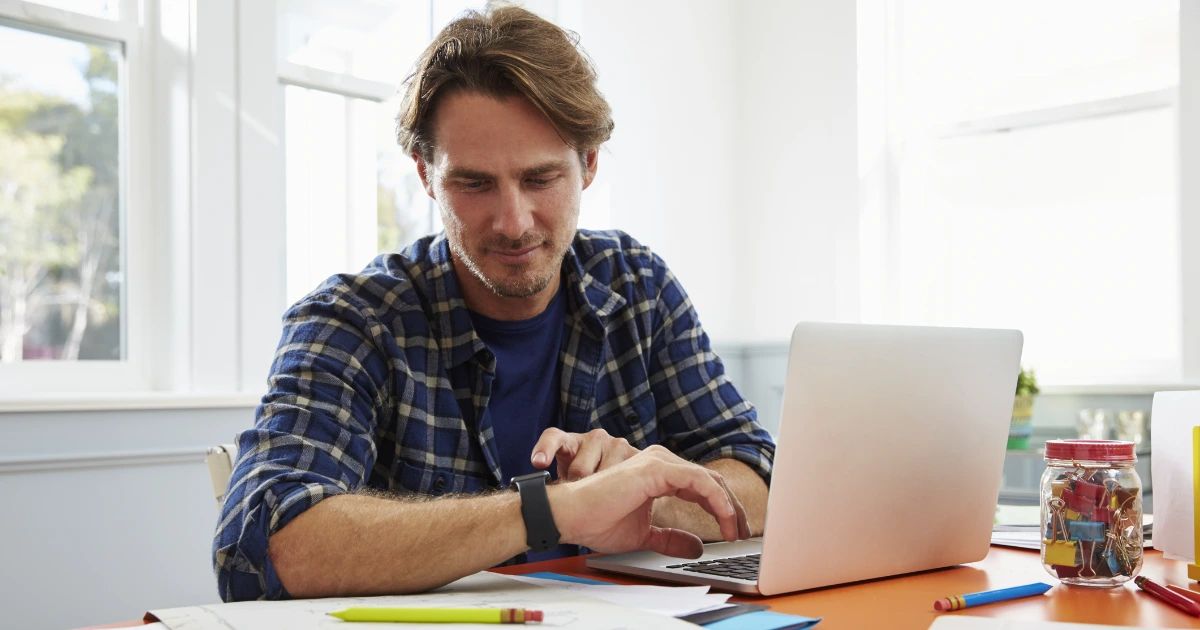 Man in plaid shirt typing on laptop at bright home office desk with pencils and paper clips nearby