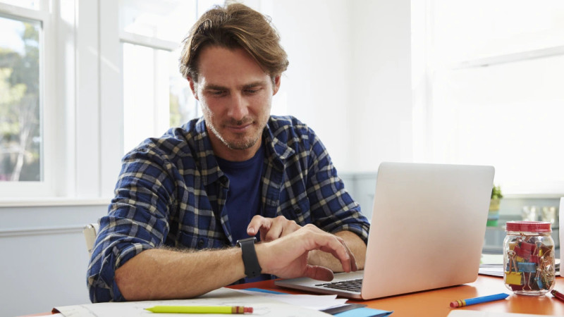 Man in plaid shirt typing on laptop at bright home office desk with pencils and paper clips nearby