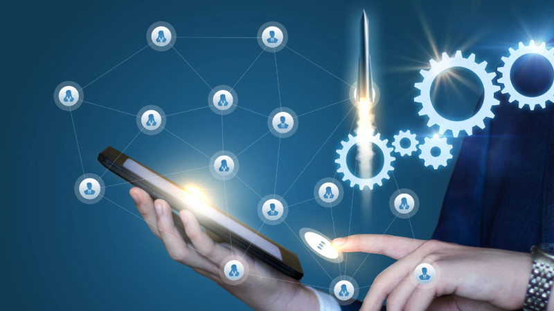 Suited person holding smartphone with glowing digital network icons, rocket and gear graphics overlaid on blue background