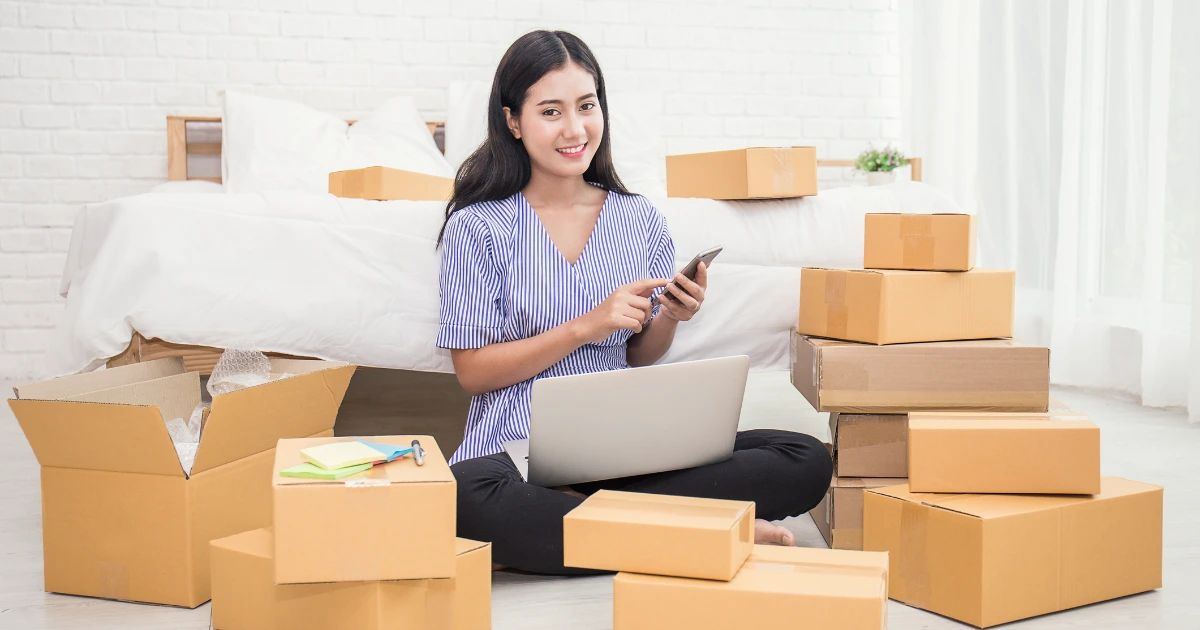 Young woman sitting on floor surrounded by cardboard boxes, using laptop and smartphone in a home bedroom setting