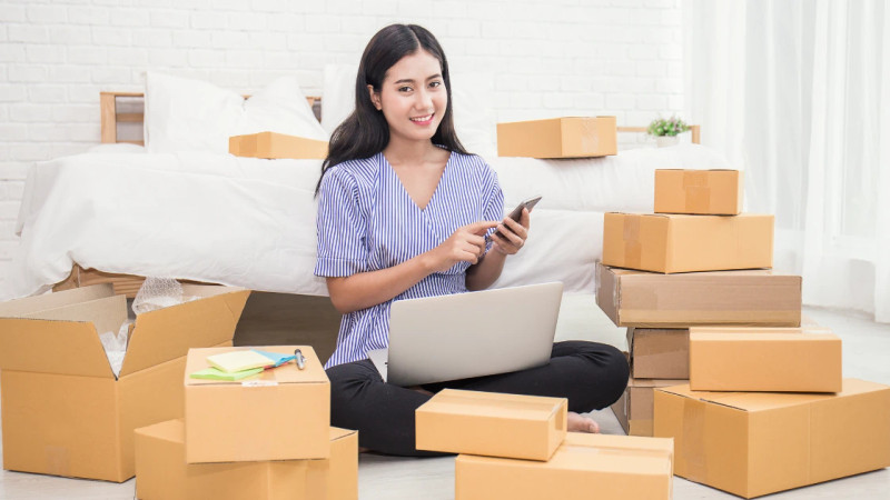 Young woman sitting on floor surrounded by cardboard boxes, using laptop and smartphone in a home bedroom setting