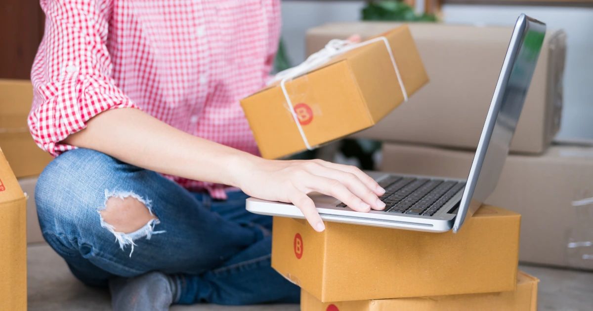 Person in checked shirt holding parcel box while typing on laptop, surrounded by cardboard shipping boxes