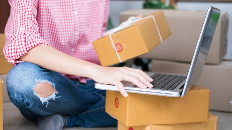 Person in checked shirt holding parcel box while typing on laptop, surrounded by cardboard shipping boxes