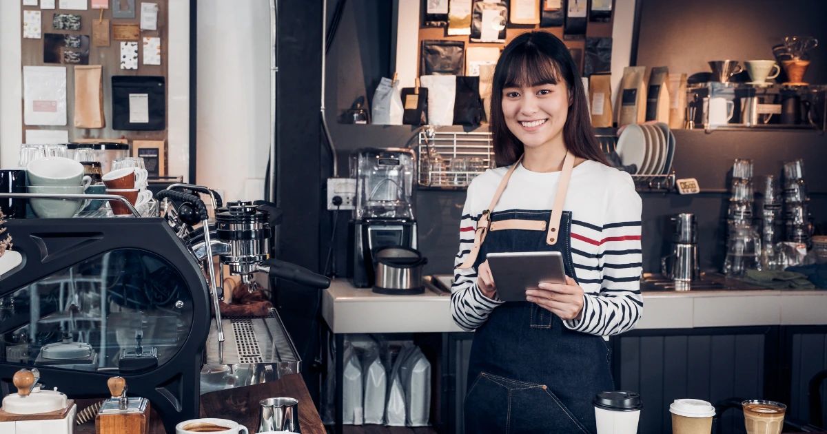 Smiling young woman in apron holding a tablet behind a coffee shop counter with espresso machine
