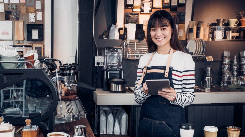 Smiling young woman in apron holding a tablet behind a coffee shop counter with espresso machine