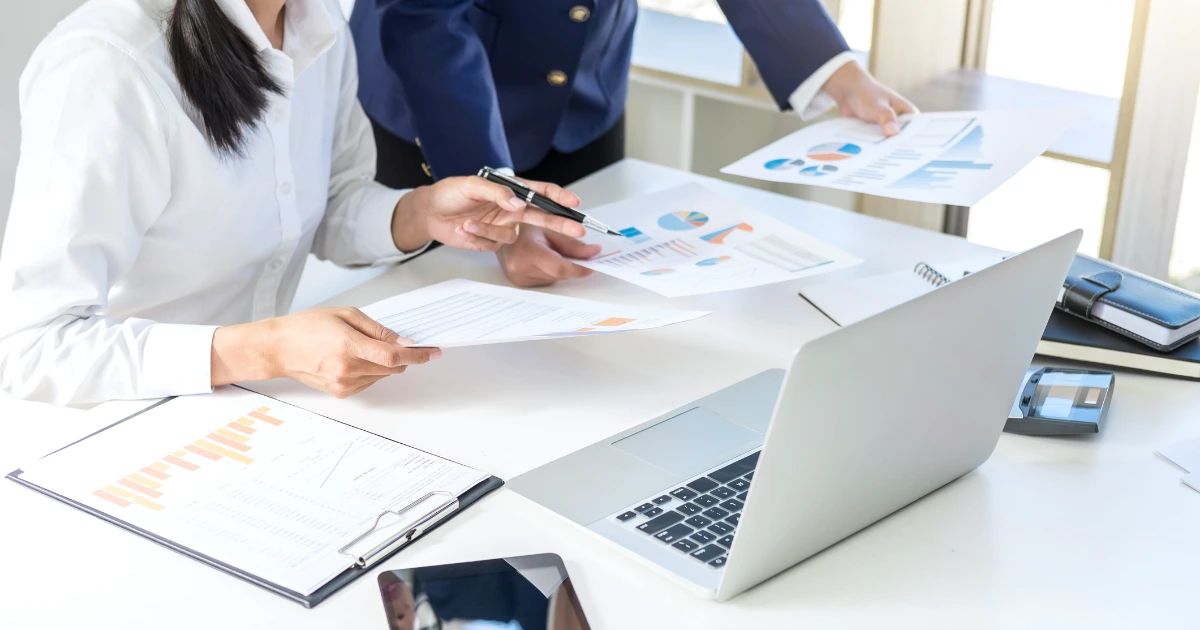 Two people at a desk reviewing printed charts and graphs, with a laptop, clipboard and tablet visible