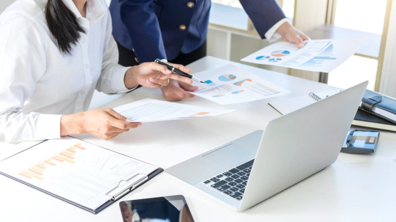 Two people at a desk reviewing printed charts and graphs, with a laptop, clipboard and tablet visible