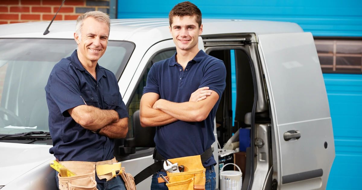 Two male tradesmen in navy workwear with tool belts standing arms crossed beside a white van and blue roller door