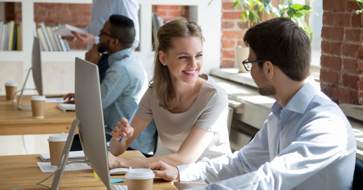 Two colleagues talking at a desktop computer in a bright open-plan co-working space with exposed brick walls