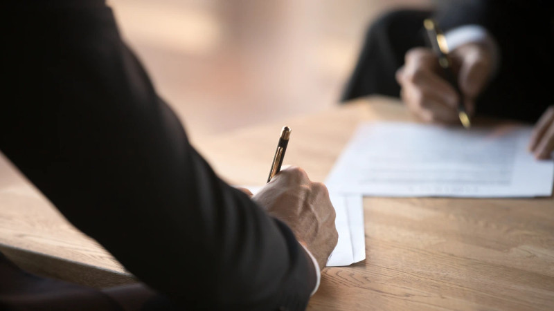 Two suited individuals signing printed documents with gold pens on a wooden table, close-up shot.