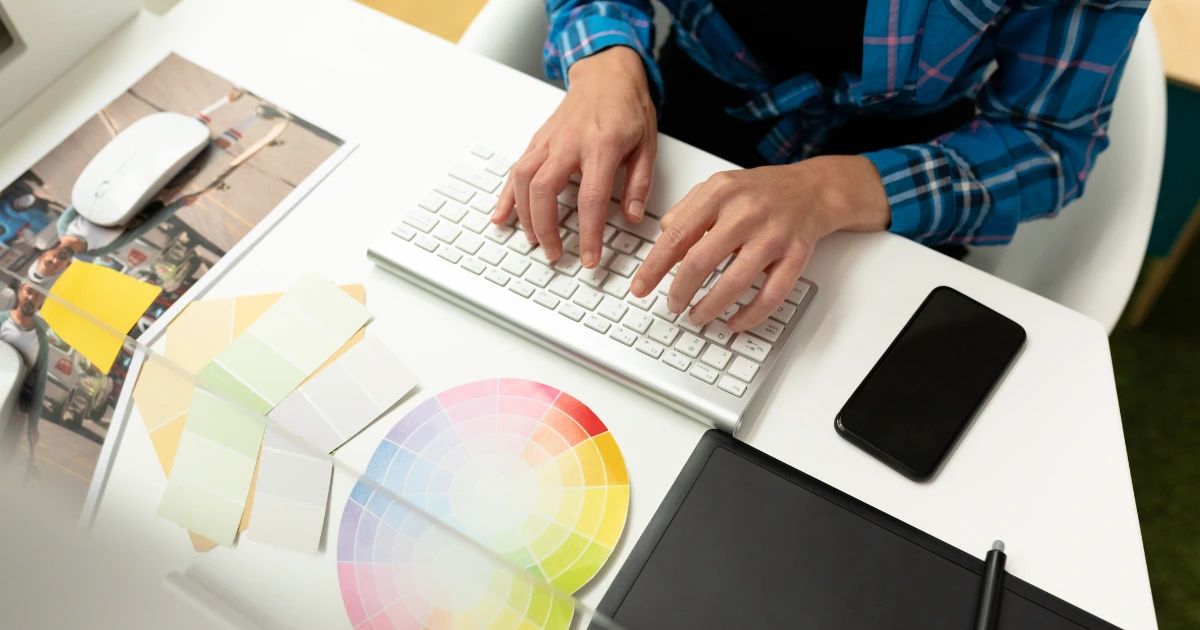 Person in plaid shirt typing at keyboard surrounded by colour swatches, graphics tablet, phone and printed photos