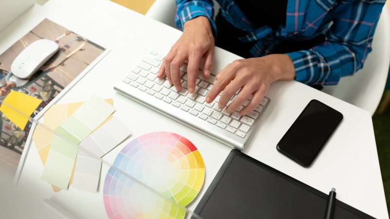 Person in plaid shirt typing at keyboard surrounded by colour swatches, graphics tablet, phone and printed photos