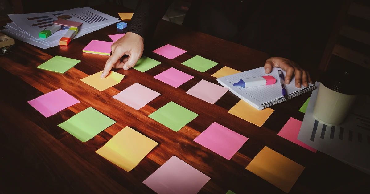Two pairs of hands arranging colourful sticky notes in a grid on a dark wooden desk with a notepad and highlighters