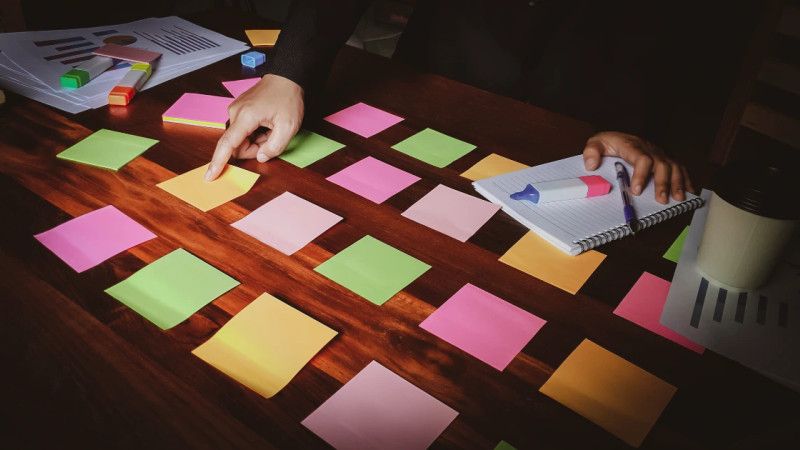Two pairs of hands arranging colourful sticky notes in a grid on a dark wooden desk with a notepad and highlighters