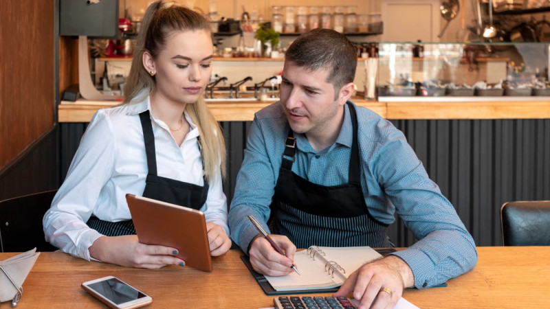 Two café owners in black aprons review a tablet and notebook with calculator at a wooden counter