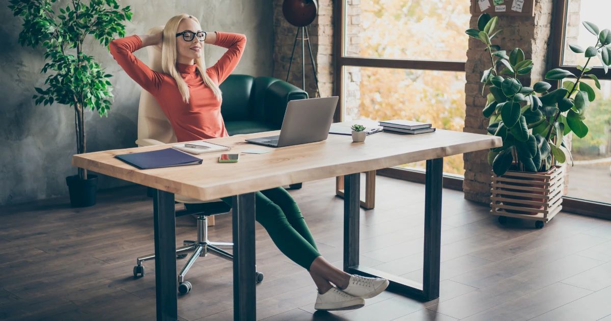 Woman with glasses leaning back in chair at wooden desk with laptop, in a loft-style office with plants