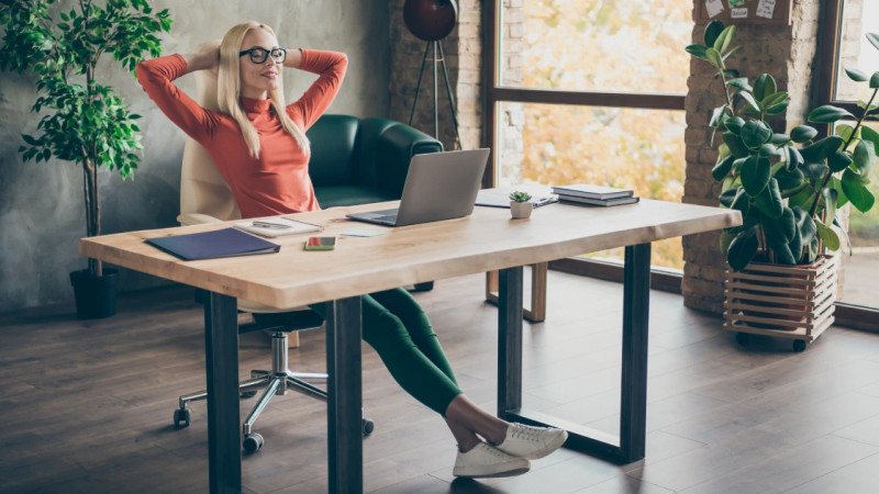 Woman with glasses leaning back in chair at wooden desk with laptop, in a loft-style office with plants