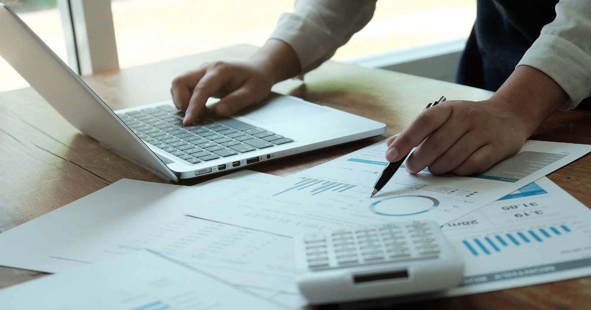 Hands at a wooden desk reviewing printed bar and pie charts, holding a pen, with a laptop and calculator nearby.