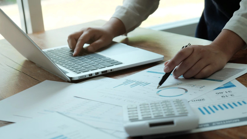 Hands at a wooden desk reviewing printed bar and pie charts, holding a pen, with a laptop and calculator nearby.