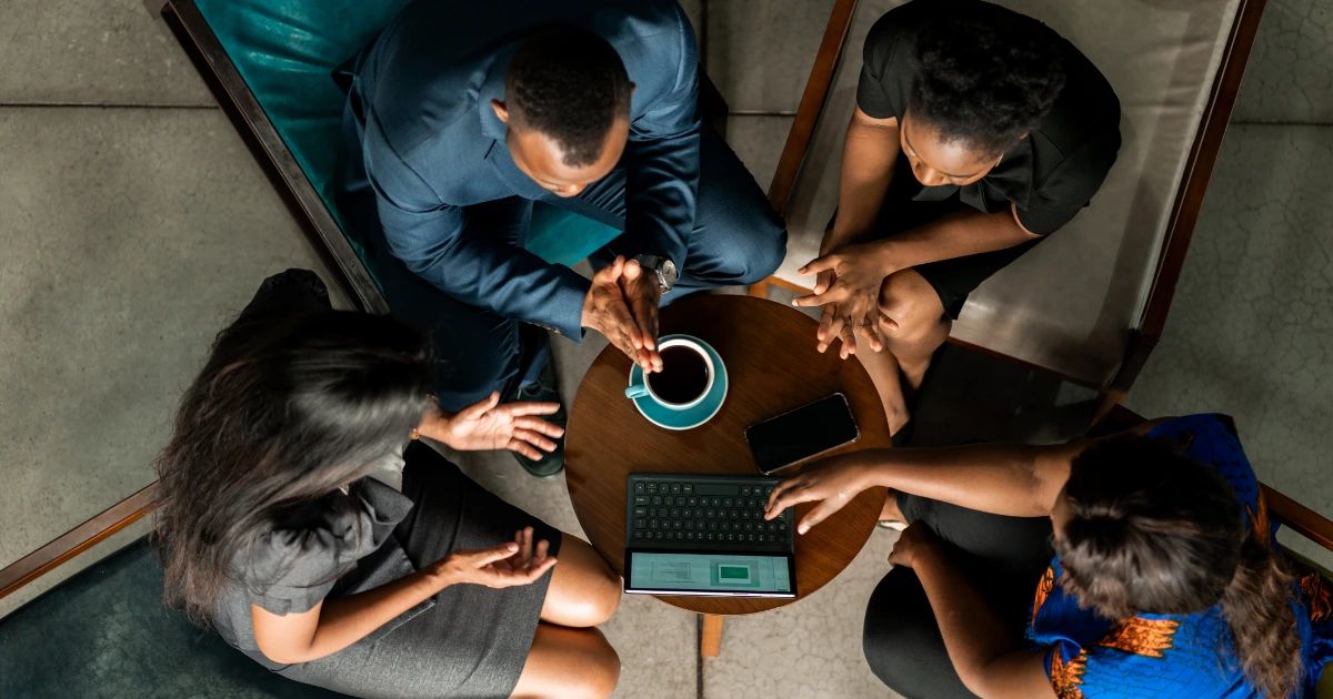 Overhead view of four people gathered around a small round table with a laptop, phone and coffee cup