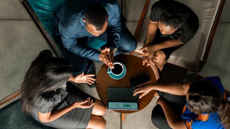 Overhead view of four people gathered around a small round table with a laptop, phone and coffee cup