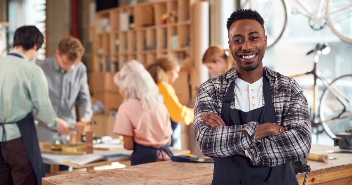 Smiling man in apron and plaid shirt stands arms crossed in a craft workshop, team working at benches behind