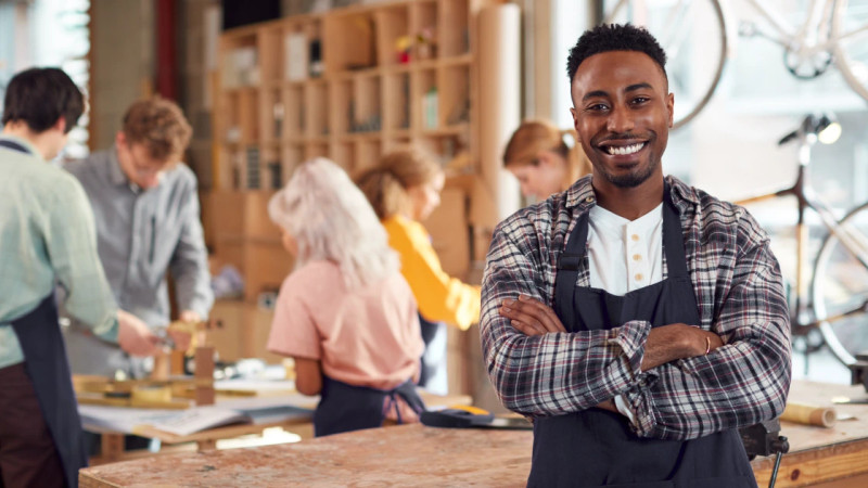 Smiling man in apron and plaid shirt stands arms crossed in a craft workshop, team working at benches behind