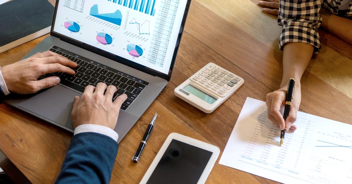 Overhead view of two people at a wooden desk with a laptop showing charts, a calculator, printed spreadsheets and a pen
