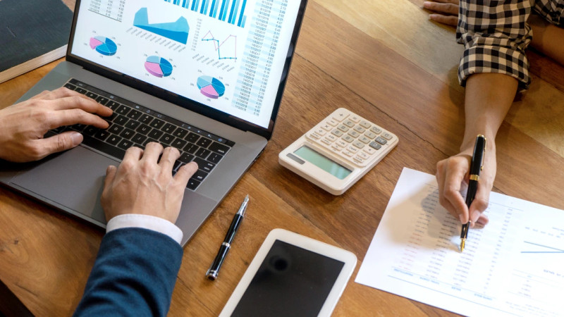 Overhead view of two people at a wooden desk with a laptop showing charts, a calculator, printed spreadsheets and a pen