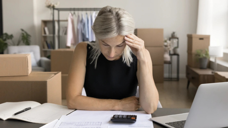 Woman with grey hair studies paperwork and calculator at desk, cardboard boxes and clothing rail behind her