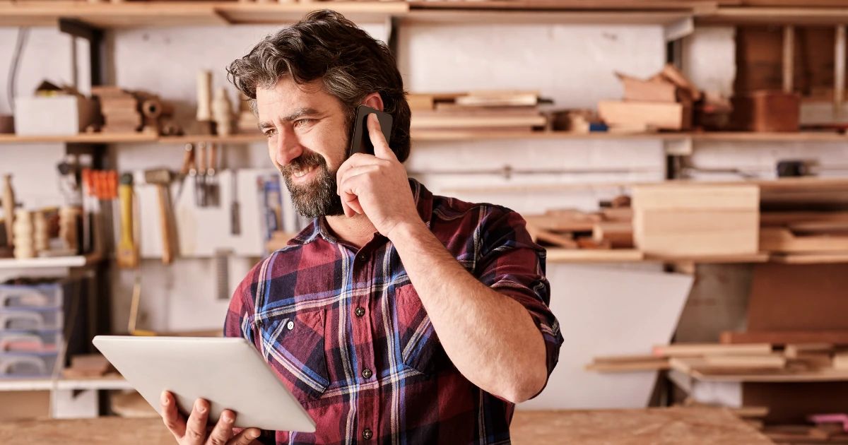 Bearded man in plaid shirt holding tablet and talking on phone in a woodworking workshop