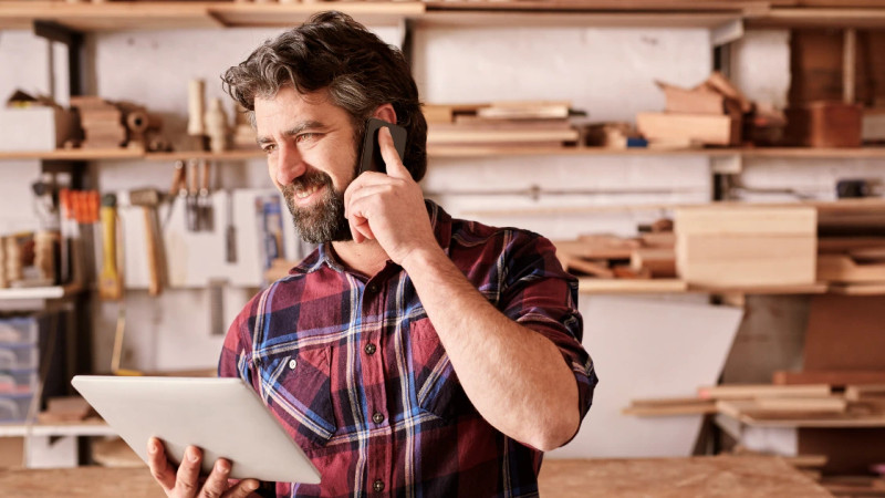 Bearded man in plaid shirt holding tablet and talking on phone in a woodworking workshop