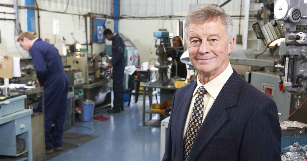 Older man in suit and tie stands in a busy engineering workshop with workers and machinery behind him