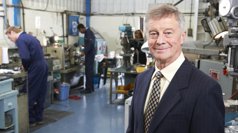 Older man in suit and tie stands in a busy engineering workshop with workers and machinery behind him