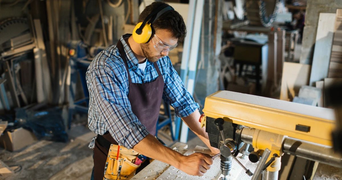 Male carpenter wearing ear defenders, safety goggles and apron operating a yellow drill press in a timber workshop