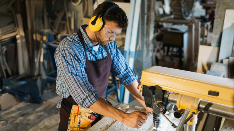 Male carpenter wearing ear defenders, safety goggles and apron operating a yellow drill press in a timber workshop