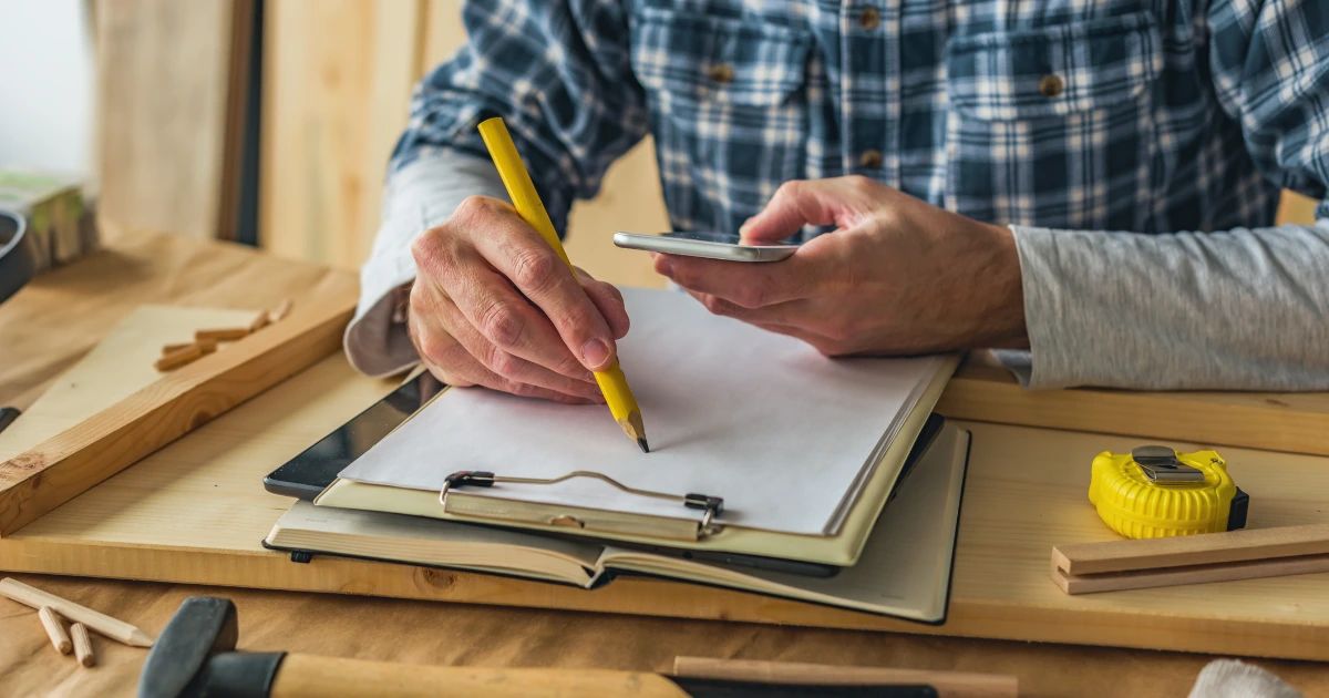 Tradesperson in plaid shirt writing on clipboard with pencil, holding smartphone, tape measure and wood on workbench